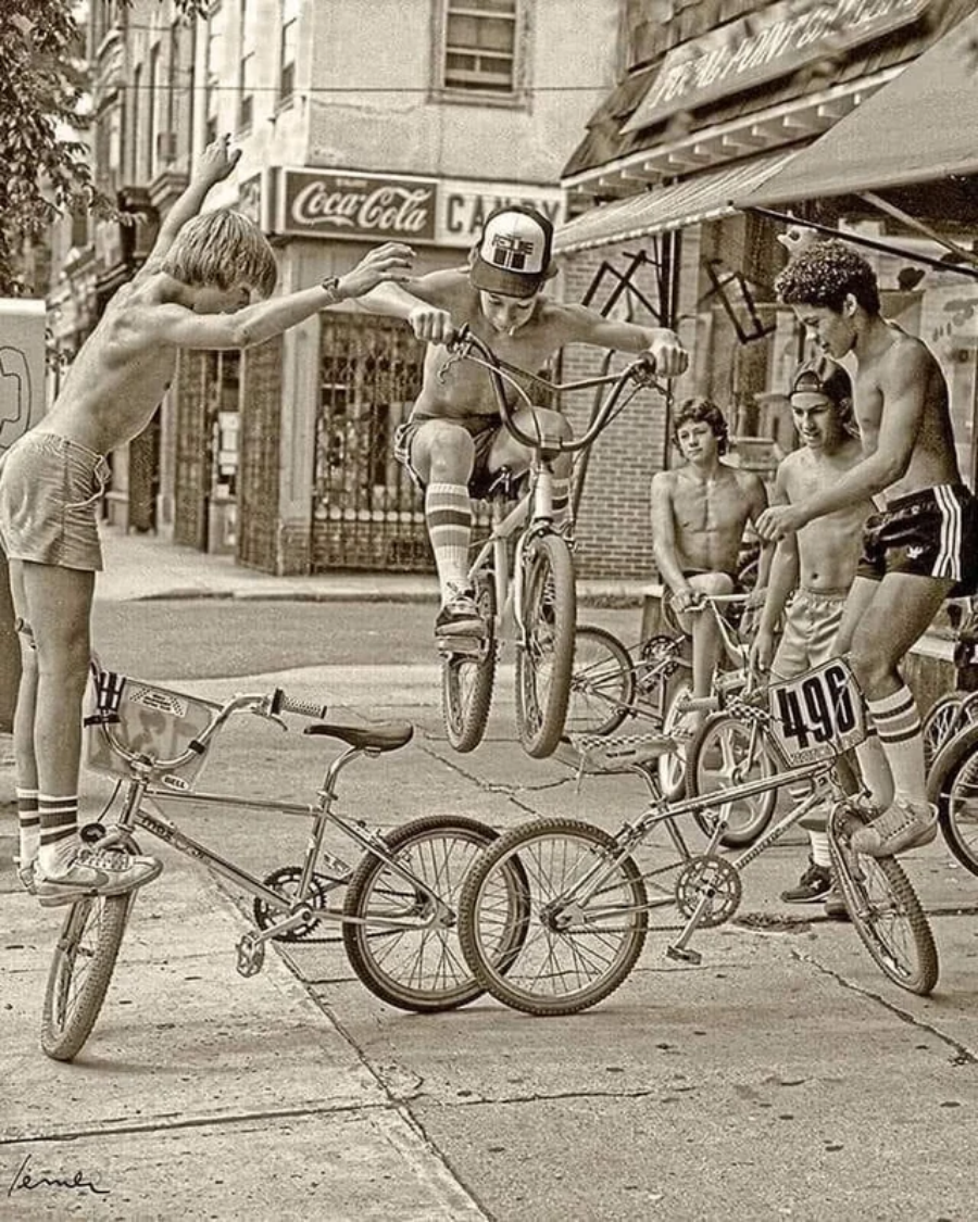 A group of boys in shorts and no shirts watch as one jumps his BMX bike over two bikes stacked on a sidewalk, while another boy leaps with arms raised beside him. A Coca-Cola sign is visible in the background.