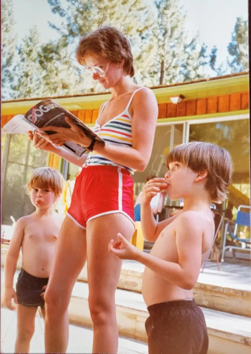 A woman in a striped tank top and red shorts reads a magazine outdoors, while two shirtless young boys stand nearby. Trees and a house with large windows are visible in the background on a sunny day.