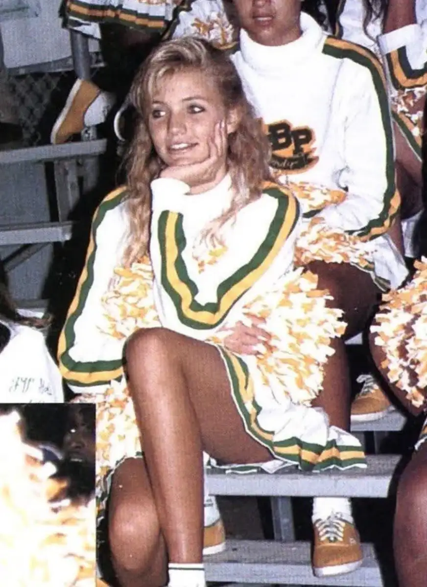 A young female cheerleader in a white and green uniform sits on bleachers, resting her chin on her hand and smiling. She holds yellow and white pom-poms, and other cheerleaders are partially visible around her.