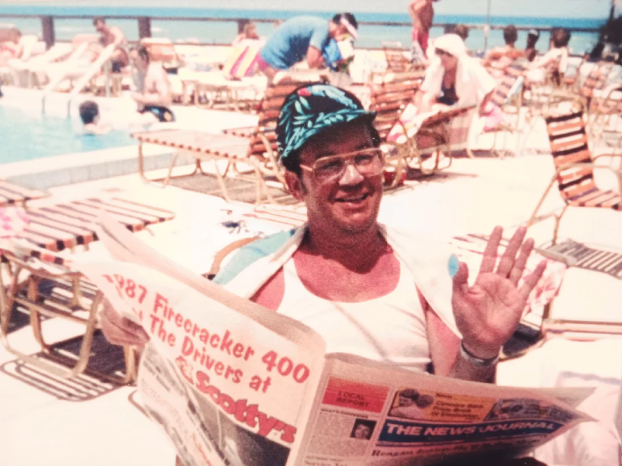 A man wearing glasses, a tropical cap, and a white tank top sits by a pool, smiling and waving while holding a newspaper. People relax on lounge chairs and swim in the background. It appears to be a sunny day.