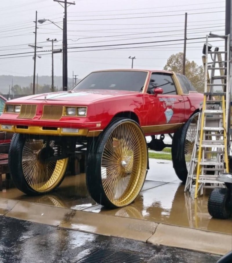 A red car with gold trim is elevated on extremely large, shiny gold wheels, parked on wet pavement near a ladder and utility poles on a rainy day.
