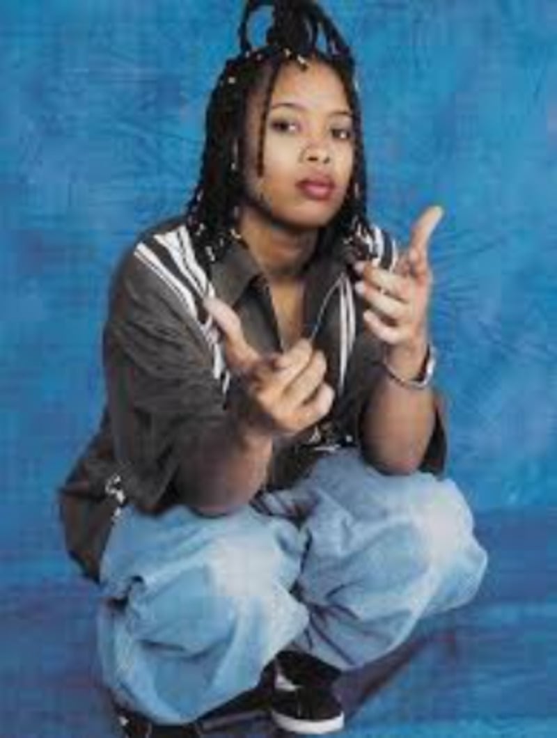 A person with braided hair squats against a blue backdrop, wearing a gray jacket and baggy jeans, looking at the camera with both hands raised in a gesture.