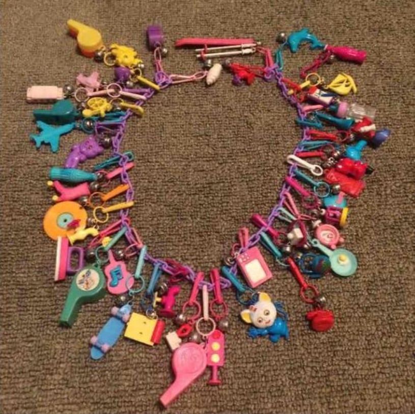 A colorful toy charm necklace laid on a brown carpet, featuring various small plastic charms like animals, whistles, musical notes, hearts, and cartoon characters attached to a bright chain.