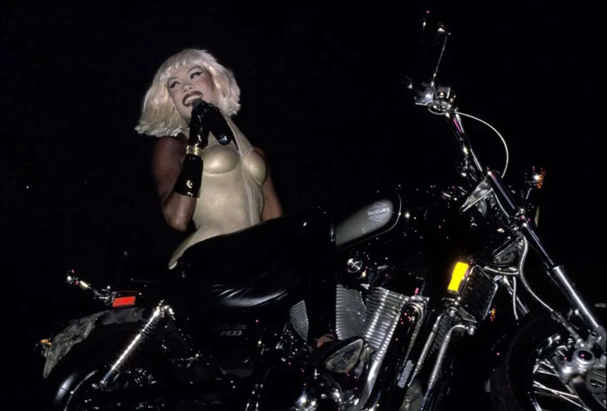 A person with platinum blonde hair, wearing a gold bodysuit and black gloves, sits on a motorcycle at night, holding a microphone and smiling while performing.