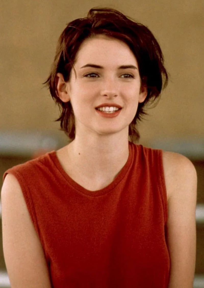 A young woman with short, dark hair smiles softly while wearing a sleeveless red top. The background is blurred and neutral, drawing focus to her face.