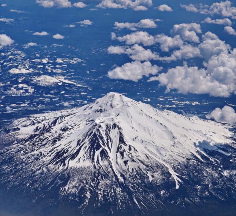 A snow-capped mountain peak, surrounded by scattered clouds and a dark forest landscape below, seen from above on a clear day.