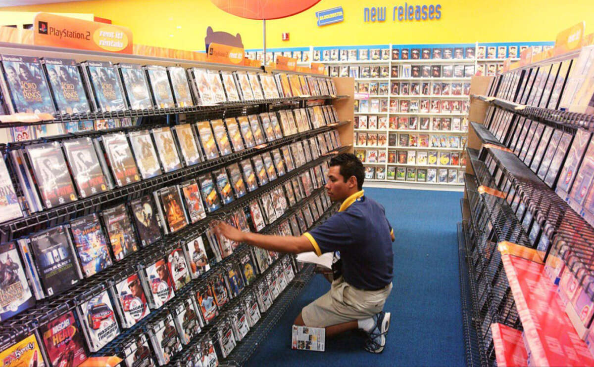 A store employee in a yellow-trimmed uniform kneels in an aisle, stocking or arranging DVD or game cases on shelves, with rows of cases and a "new releases" sign visible in the brightly lit store.