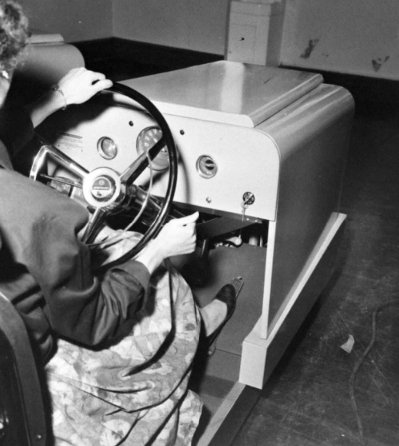 A person in a skirt sits at a vintage car driving simulator, holding the steering wheel and operating the pedals. The simulator features a dashboard with gauges and a flat metal front.
