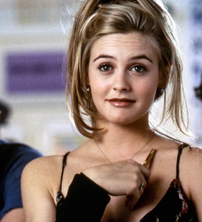 A young woman with blonde hair in a high ponytail looks slightly amused, wearing a black strappy top and holding a pen near her chest. She appears to be indoors, with a blurred background behind her.