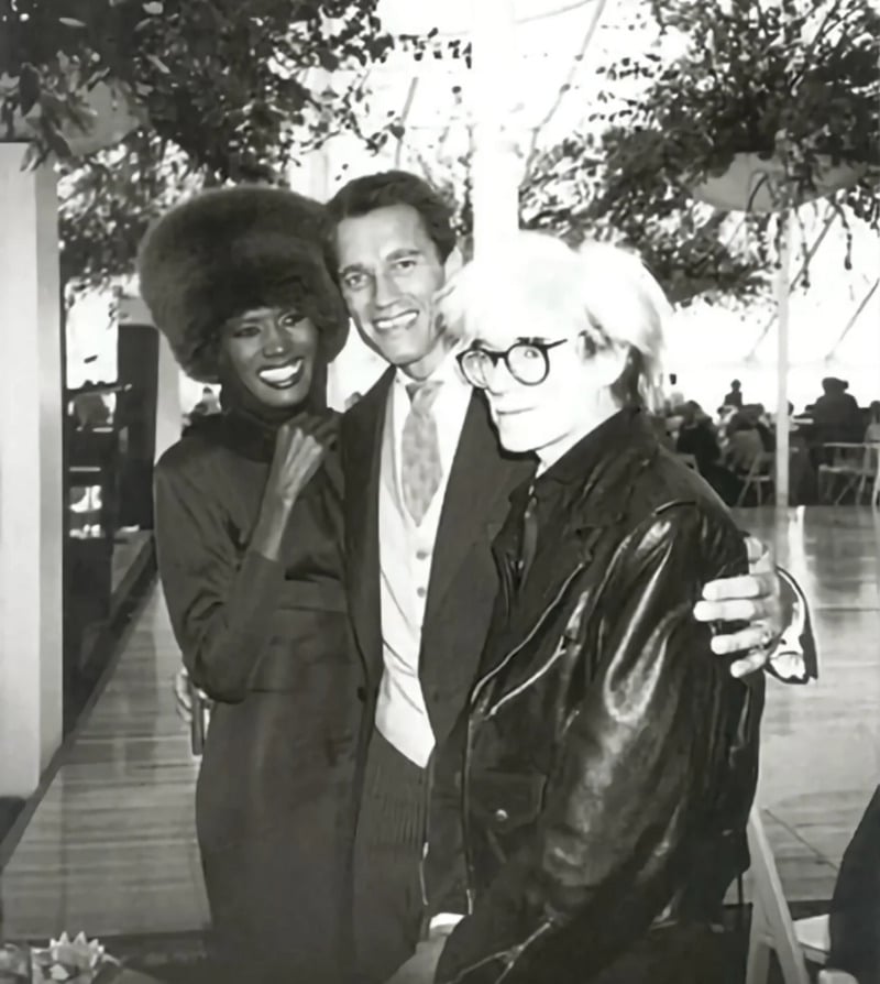 Three people pose together indoors. On the left, a woman wearing a fur hat and a dark dress smiles. In the center, a man in a suit smiles with his arms around the others. On the right, a person in glasses and a leather jacket looks at the camera.