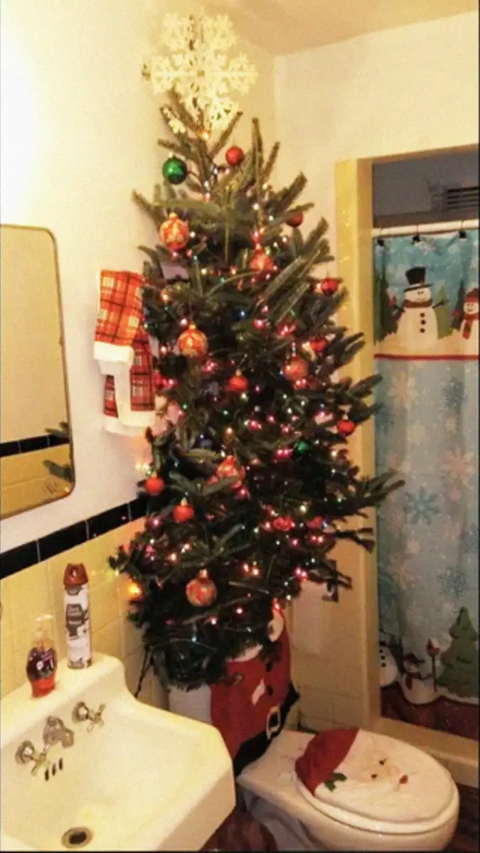 A decorated Christmas tree is positioned in a small bathroom, rising above the toilet. The bathroom features holiday-themed decorations, including a Santa seat cover and a snowman shower curtain.