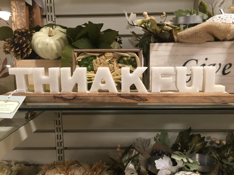 White decorative letters arranged to spell “THNAKEFUL” sit on a wooden base, surrounded by autumn-themed decor, greenery, and a small white pumpkin on a shelf.
