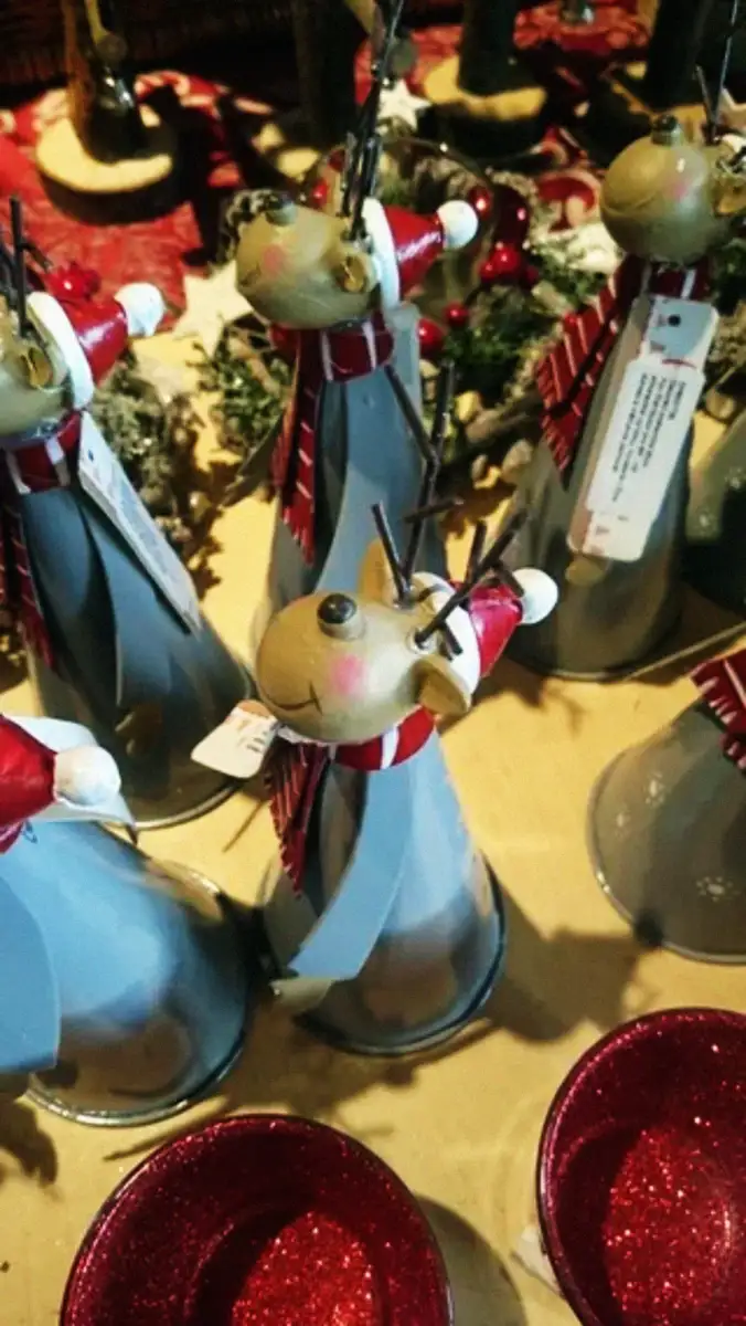 Metal cone-shaped reindeer figurines with red scarves and Santa hats are arranged on a table with red glittery bowls in the foreground. The reindeer have smiling faces and twig antlers.