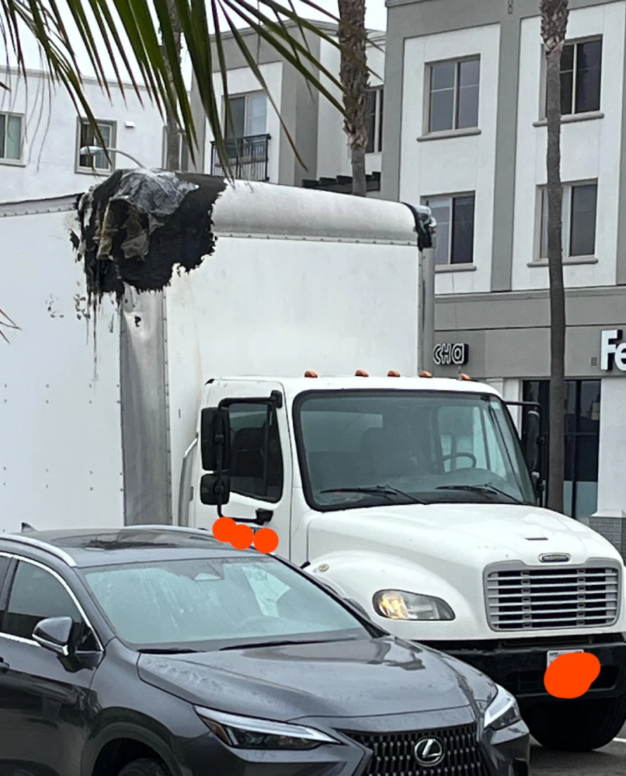 A white box truck with fire damage on its upper front corner is parked next to a gray Lexus SUV on a city street lined with palm trees and apartment buildings.