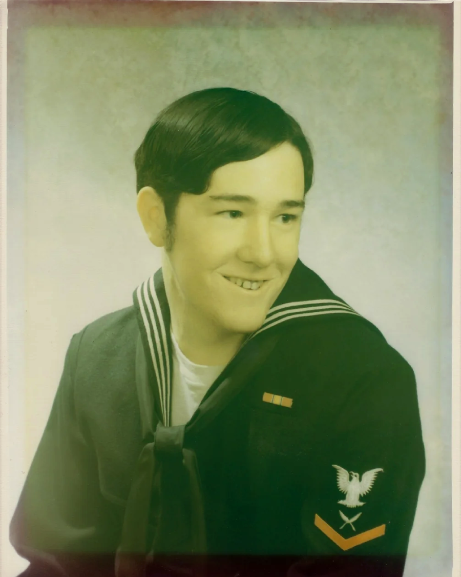A young man in a U.S. Navy uniform with dark hair sits for a formal portrait, smiling slightly. The uniform features an eagle patch with a red chevron and a ribbon above the left breast.