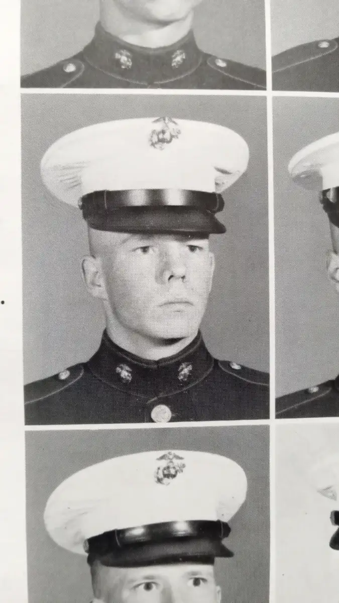 Black-and-white photo of a young Marine in uniform, including a white cap and dark jacket with buttons and insignia, looking straight ahead with a serious expression.