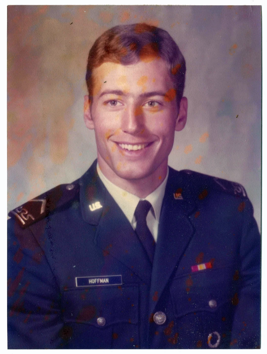 A young man in a U.S. military dress uniform smiles at the camera. His name tag reads "Hoffman." The background is plain, and there are some colored spots on the image.