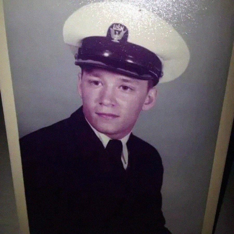 A young man in a dark navy uniform and a white hat with a USN insignia poses for a formal portrait against a plain background.