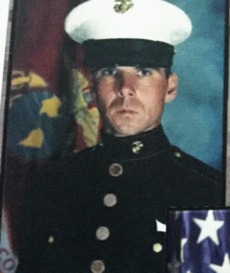 A man in a Marine Corps dress uniform stands in front of a flag with a serious expression; part of a folded American flag is visible in the lower right corner.