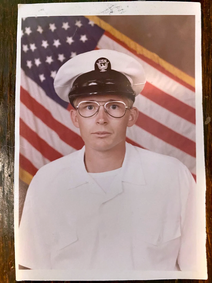 A young person in a U.S. Navy uniform and glasses poses for a portrait in front of an American flag backdrop.