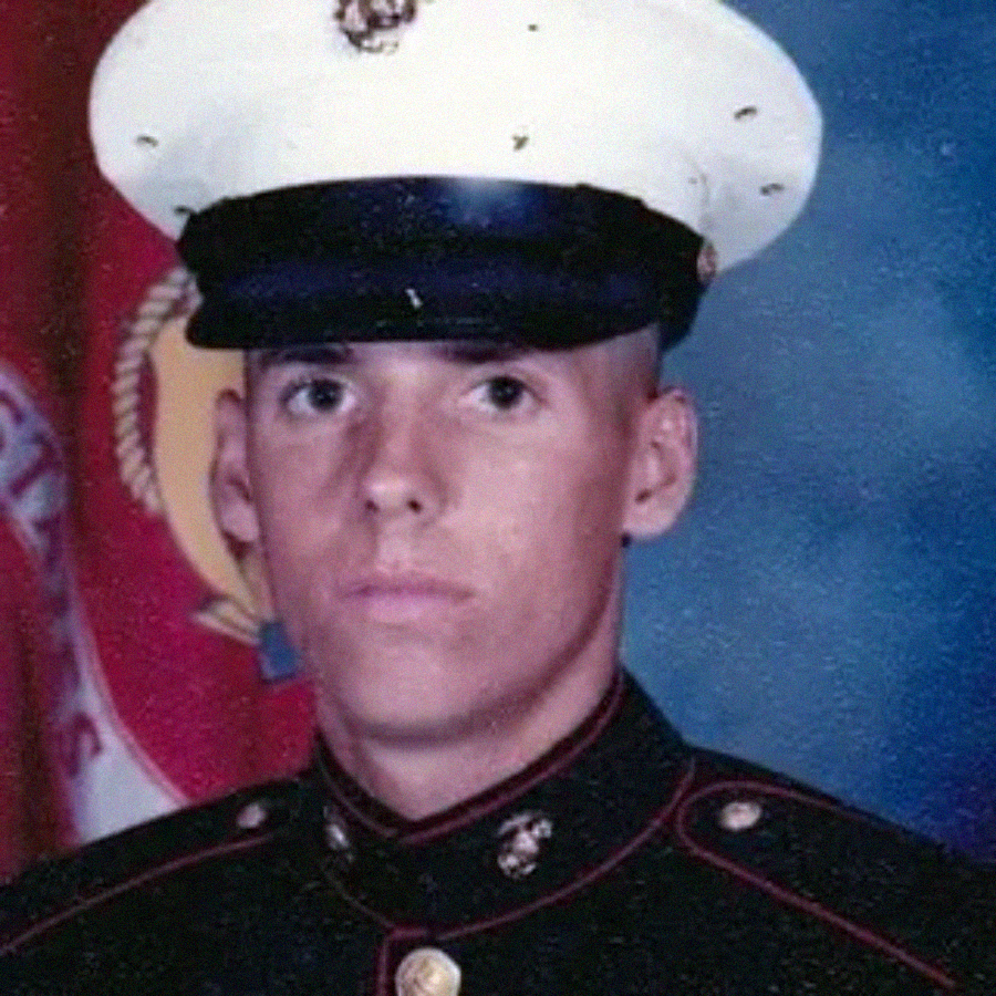 A young man in a U.S. Marine Corps dress uniform poses for a formal portrait in front of an American flag and a Marine Corps flag.