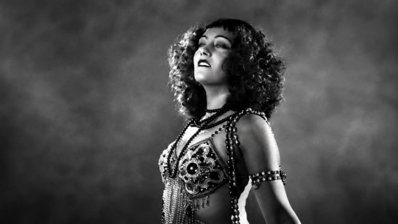 A woman with curly hair in a beaded costume poses confidently against a dark, smoky background in a black-and-white photo. 1925