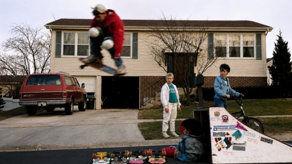 A child on a skateboard jumps off a homemade ramp while two boys watch and another sits on the curb; a red SUV and a house are in the background. 1980s