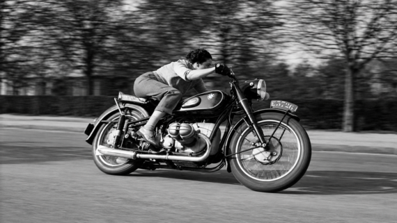 A person leans forward, riding a vintage BMW motorcycle at speed on a road with blurred trees in the background, suggesting motion.