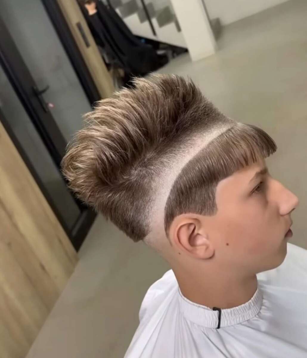 A young person with a unique haircut featuring a sharp, horizontal shaved line around the head, short bangs, and longer voluminous hair on top, sitting in a modern barbershop.