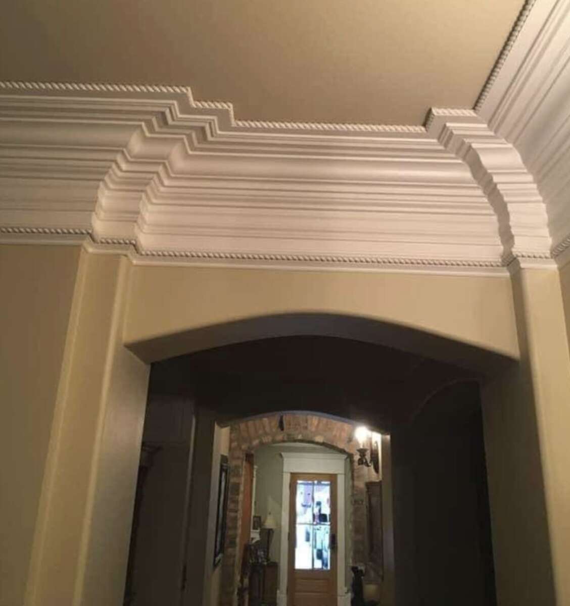 Ornate white crown molding with sharp, zigzagging layered edges contrasts with beige walls above an arched doorway in a hallway, leading to a door with glass panels and a lit chandelier in the background.