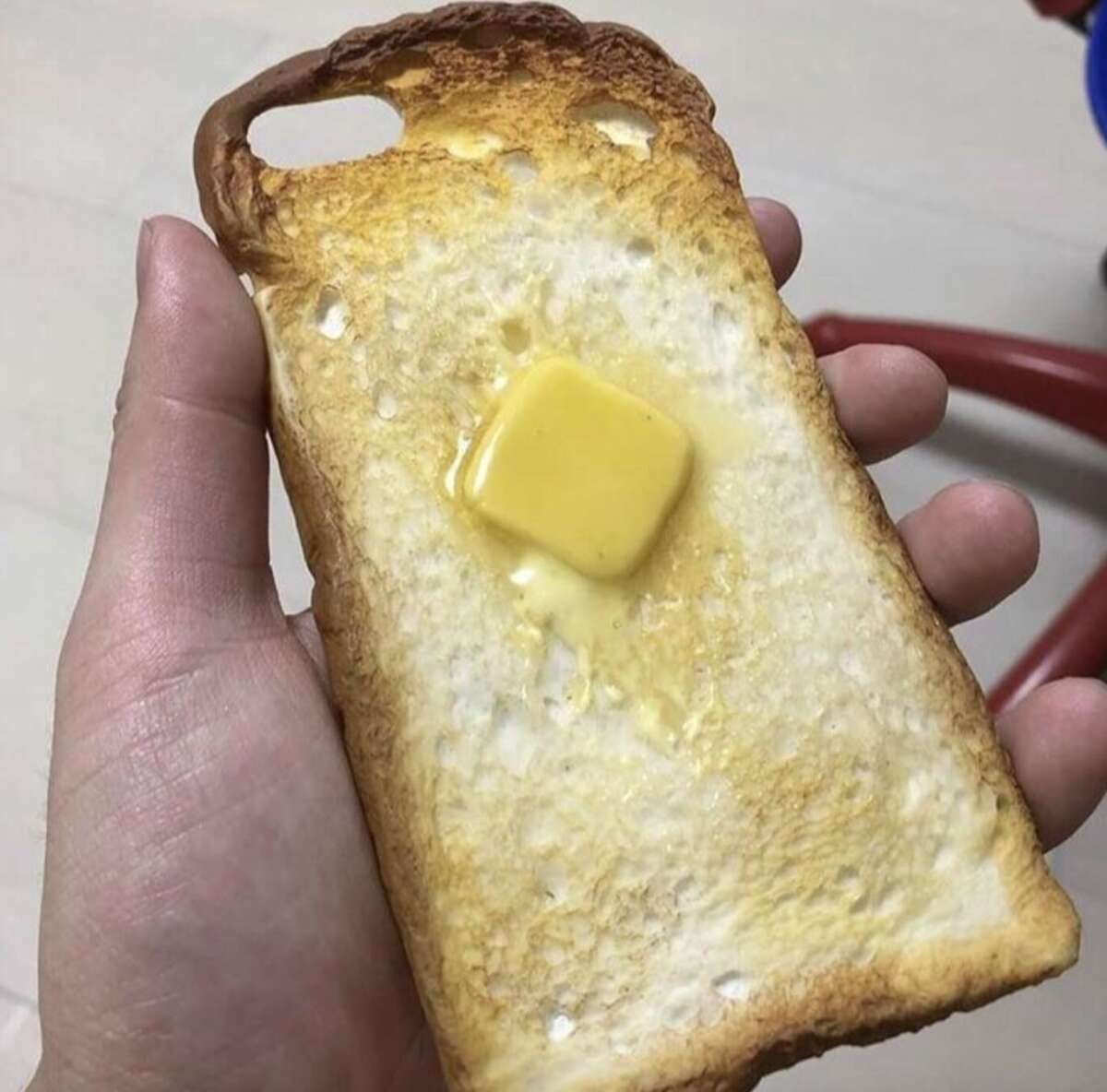 A hand holding a phone case shaped like a slice of toast with a realistic pat of melting butter in the center.