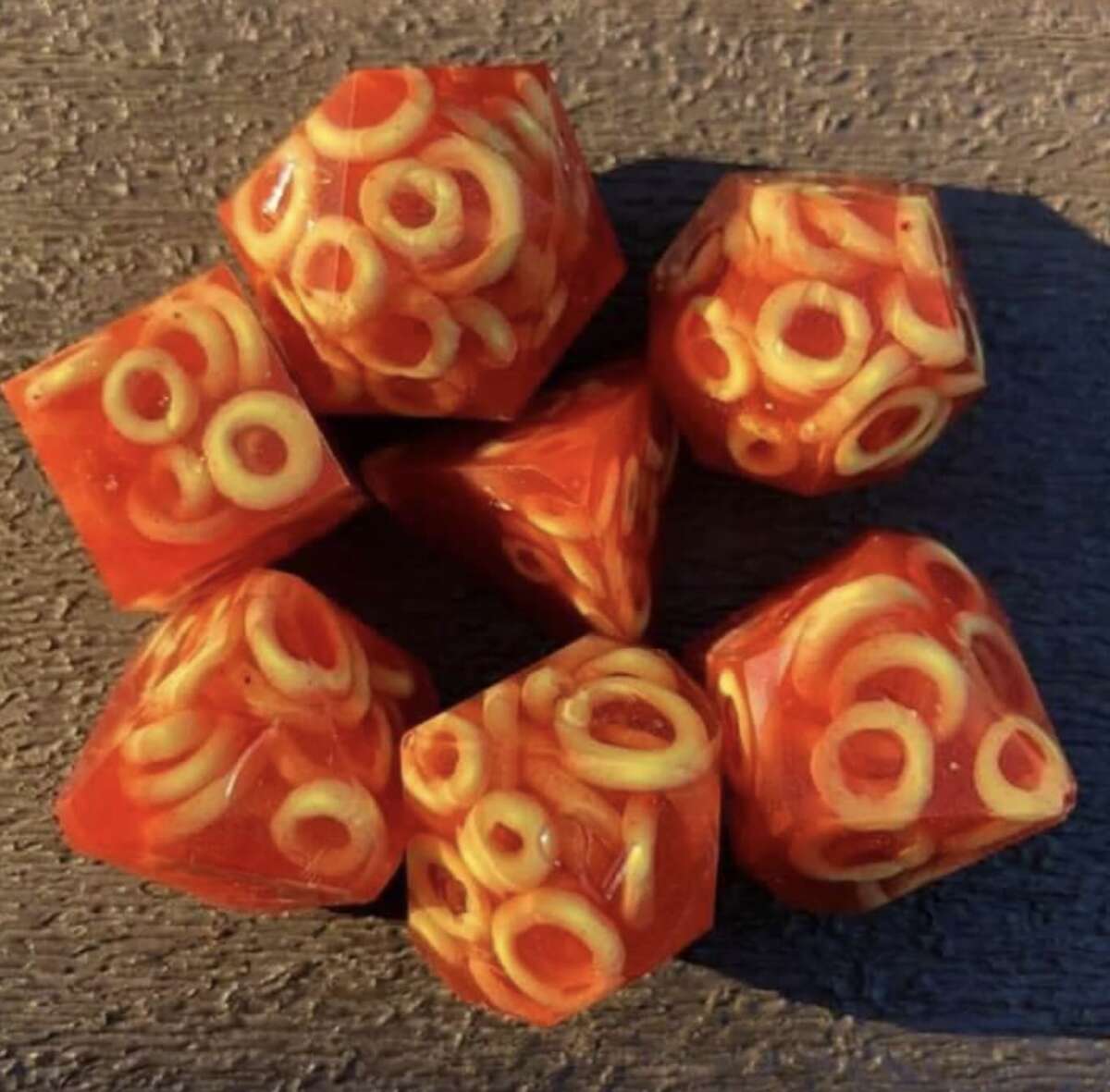 Seven polyhedral dice made of clear resin with small pasta loops and red sauce embedded inside, arranged on a textured brown surface.