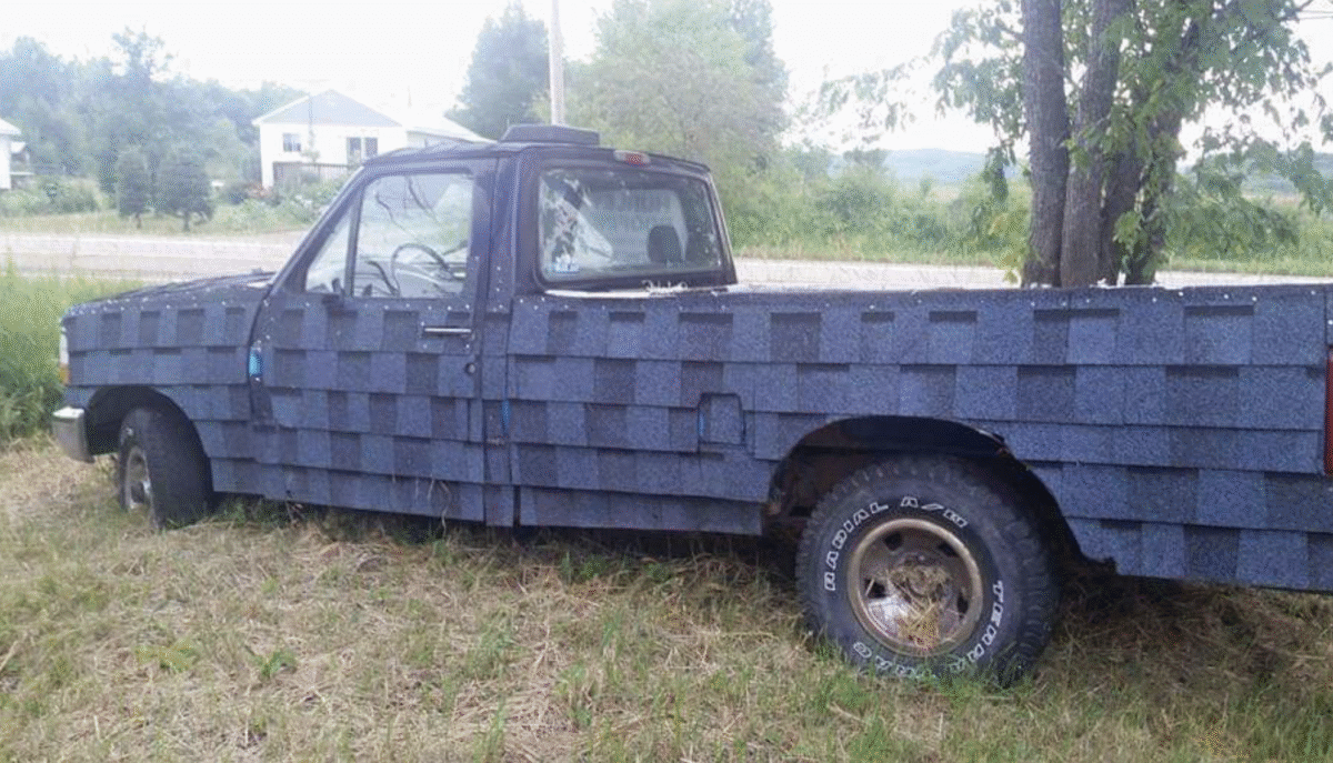 A pickup truck parked on grass is covered entirely with gray roof shingles, including the doors and body panels. Trees and a house are visible in the background.