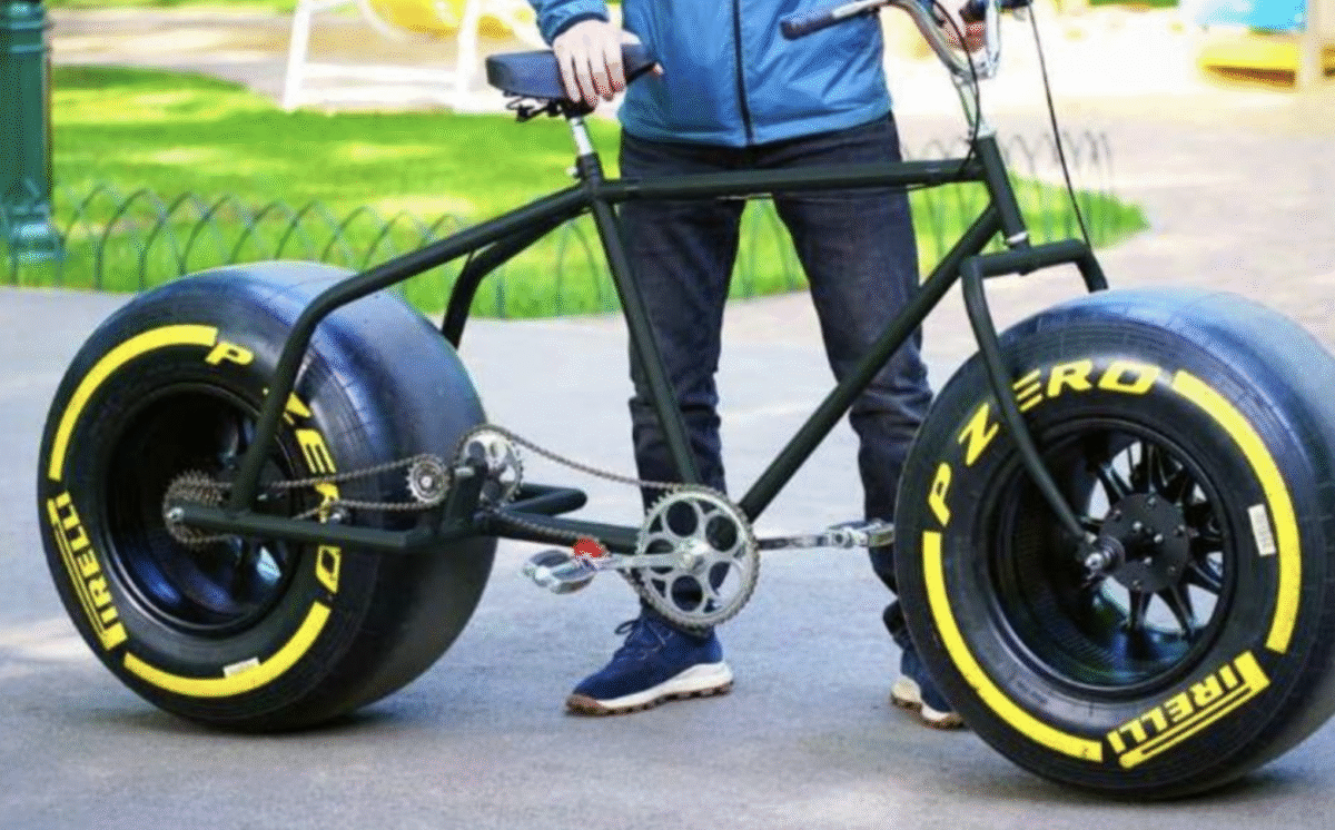 A person stands behind a large black bicycle with oversized Pirelli P Zero tires, typically used for race cars. The bike is on a paved path in a park.