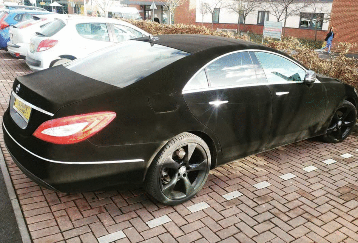 A black luxury sedan with matte, possibly velvet or suede-like exterior, parked in a brick parking lot next to other cars, with buildings and bushes in the background.