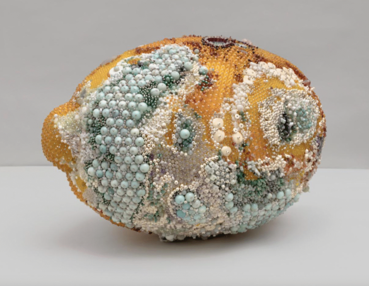 A sculpture of a lemon covered with variously sized beads and pearls in pastel colors, creating a textured and ornate surface. The background is plain and light gray.