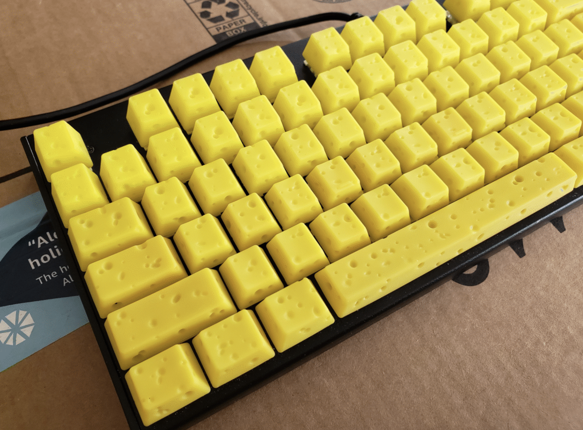 A computer keyboard with bright yellow keycaps designed to look like blocks of Swiss cheese, showing holes and uneven surfaces, placed on a cardboard box.