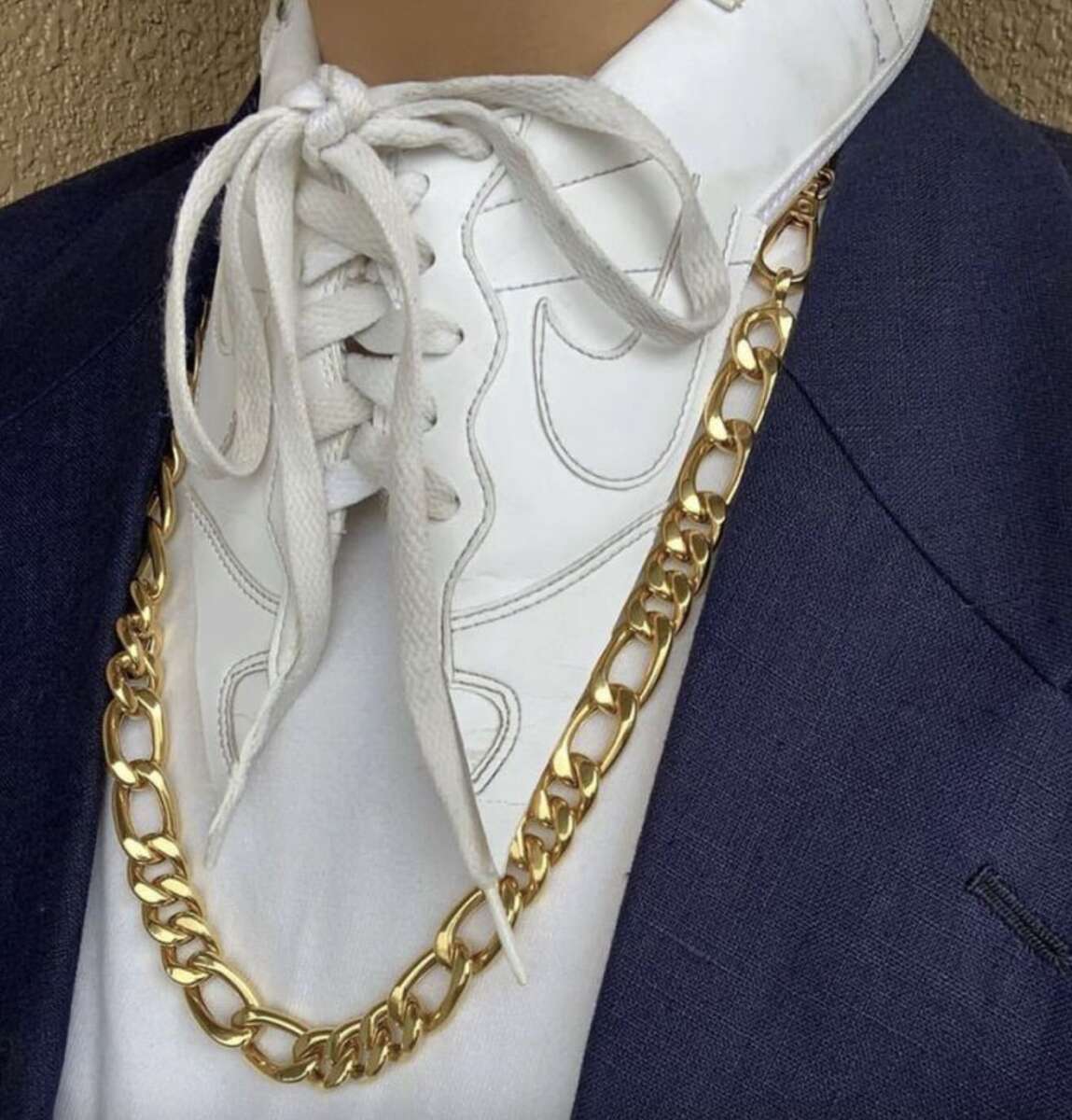 A person wears a navy suit jacket and white shirt with a sneaker tongue and laces styled as a collar, finished with a thick gold chain necklace.