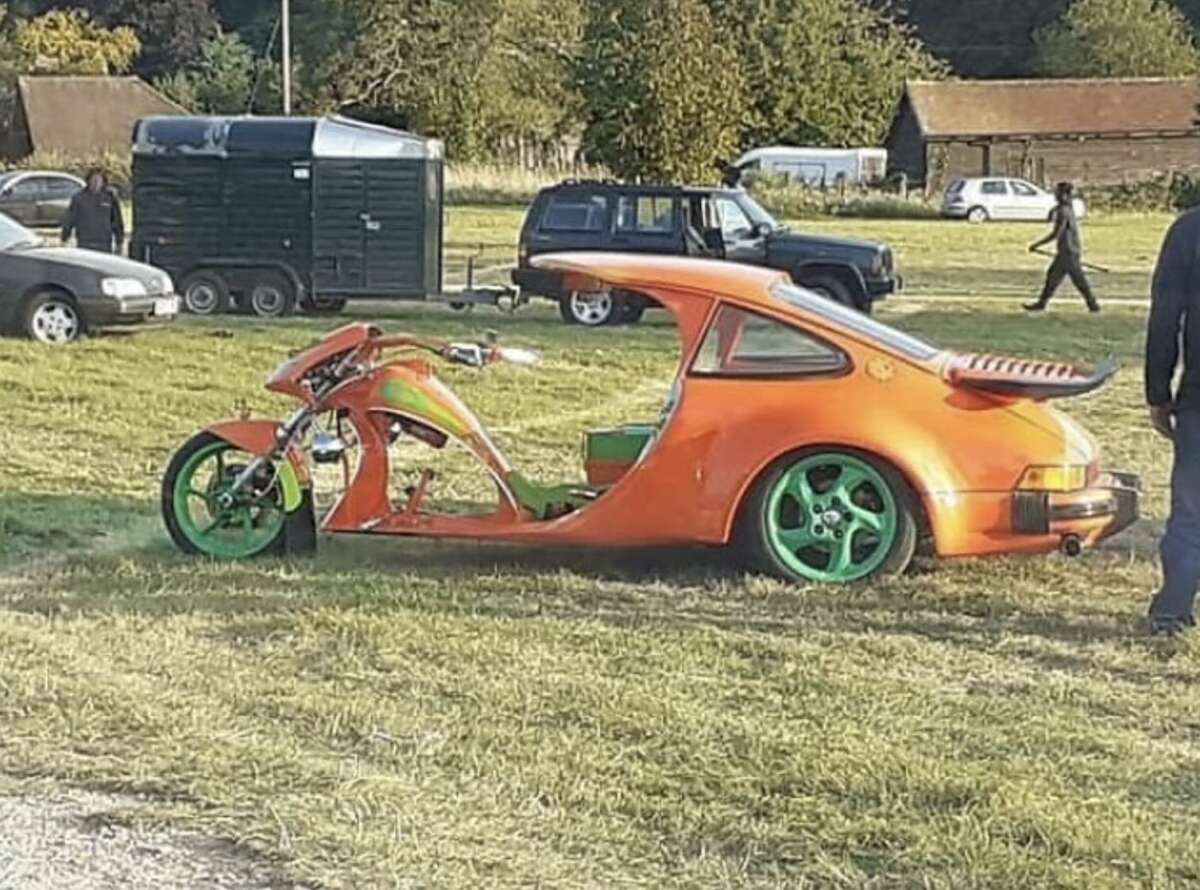 A unique vehicle combining the front of a motorcycle with the rear of an orange sports car, featuring green wheels, is parked on grass at an outdoor event with other cars and buildings in the background.