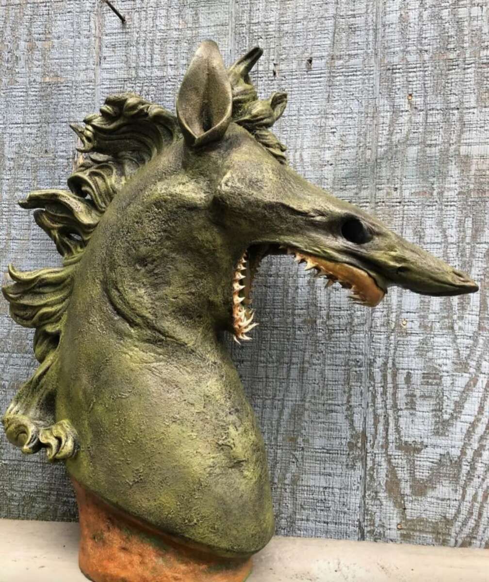 A sculpture of a mythical creature resembling a horse with an elongated snout, sharp teeth, and textured greenish skin, set against a weathered wooden background.