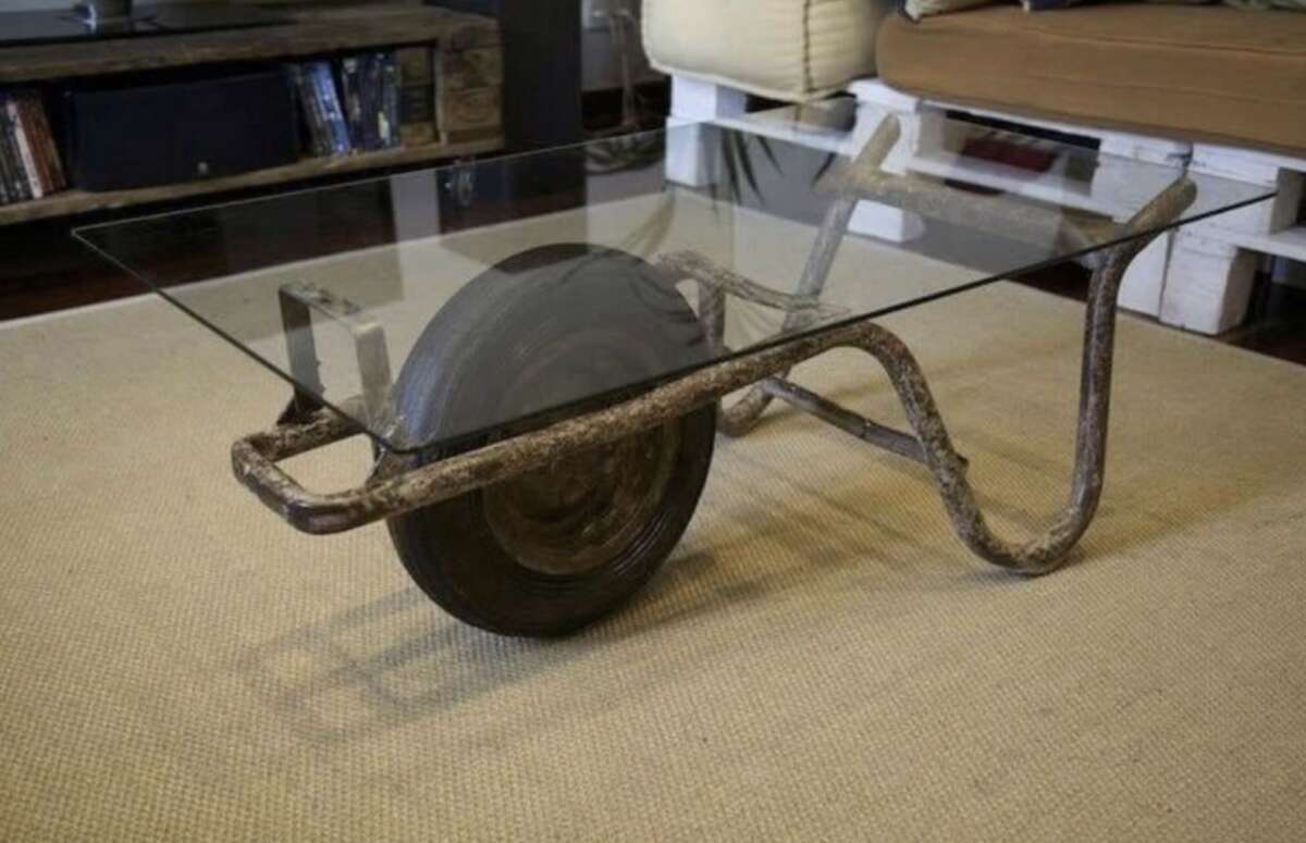 A coffee table made from an old metal wheelbarrow with a glass top, placed on a beige rug in a living room.