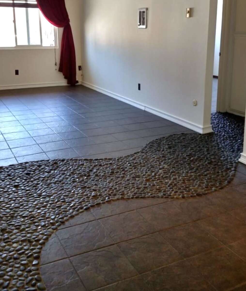 A living room with a unique floor design featuring smooth, dark river stones flowing into brown tile, creating a river-like effect between the hallway and main area; a window with red curtains is in the background.