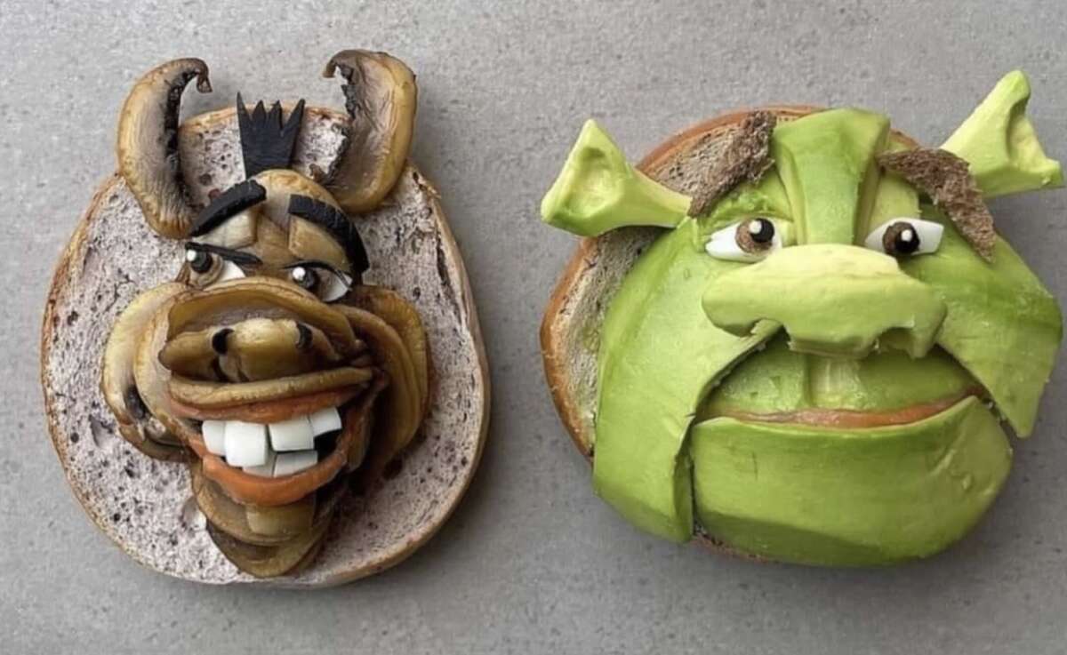 Two pieces of bread topped with vegetables and spreads arranged to resemble Donkey and Shrek from the animated movie Shrek, set on a gray surface.