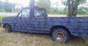 A pickup truck parked on grass, covered entirely with overlapping gray roof shingles instead of traditional paint or metal panels. Trees and a house are visible in the background.