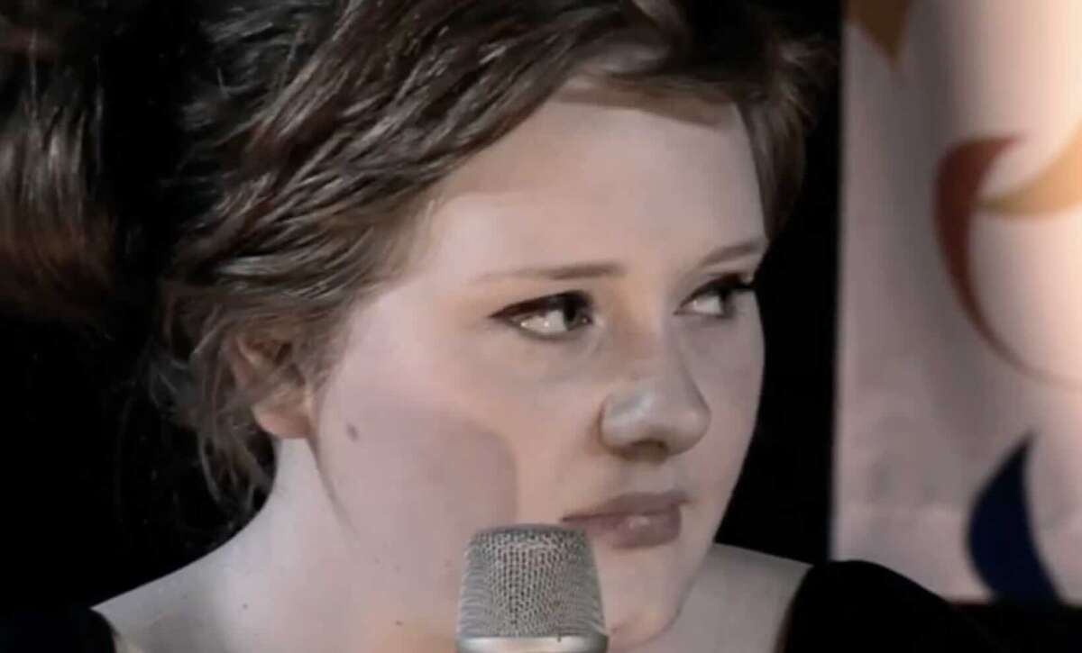 A woman with light skin and brown hair pulled back holds a silver microphone, looking slightly to the side with a serious expression. The background is dark with a partially visible blurred banner.