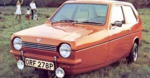 A small, orange three-wheeled car is parked on grass. In the background, a family with children and a dog enjoy a picnic outdoors. The car's license plate reads "ORF 278P.