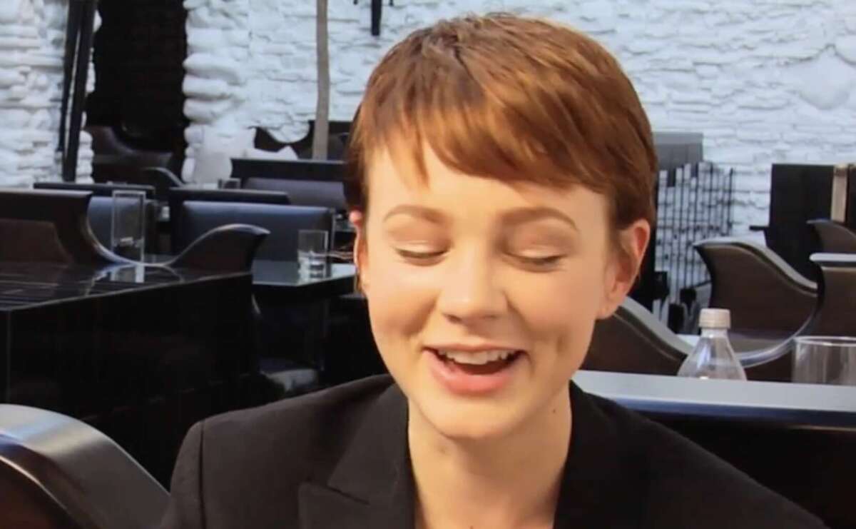 A person with short reddish-brown hair smiles with closed eyes, sitting in a modern restaurant with dark furniture and white stone walls.