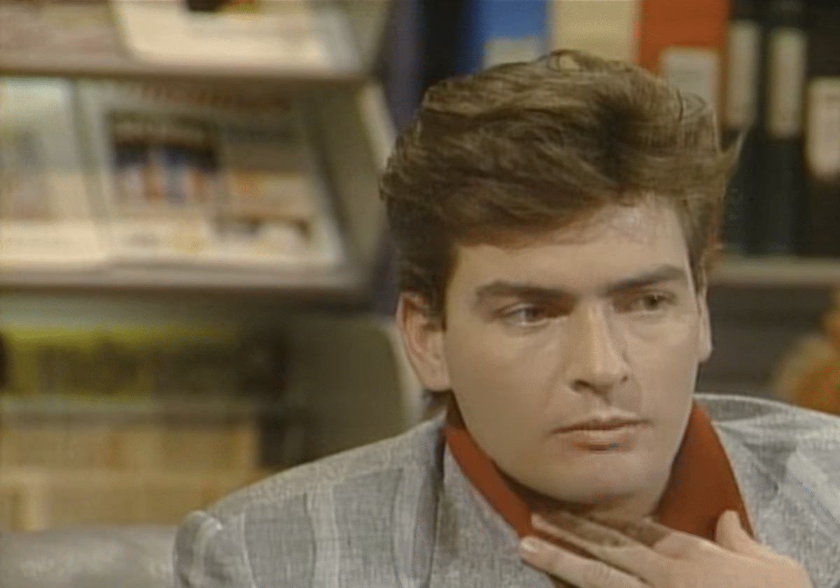 A man with brown hair, wearing a gray blazer and a red shirt, sits indoors with his hand near his neck, appearing thoughtful. Shelves with books and magazines are blurred in the background.