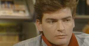A young man with short brown hair and a serious expression looks slightly to the side, wearing a gray jacket and a red shirt. Bookshelves are blurred in the background.