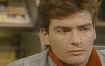 A young man with short brown hair and a serious expression looks slightly to the side, wearing a gray jacket and a red shirt. Bookshelves are blurred in the background.